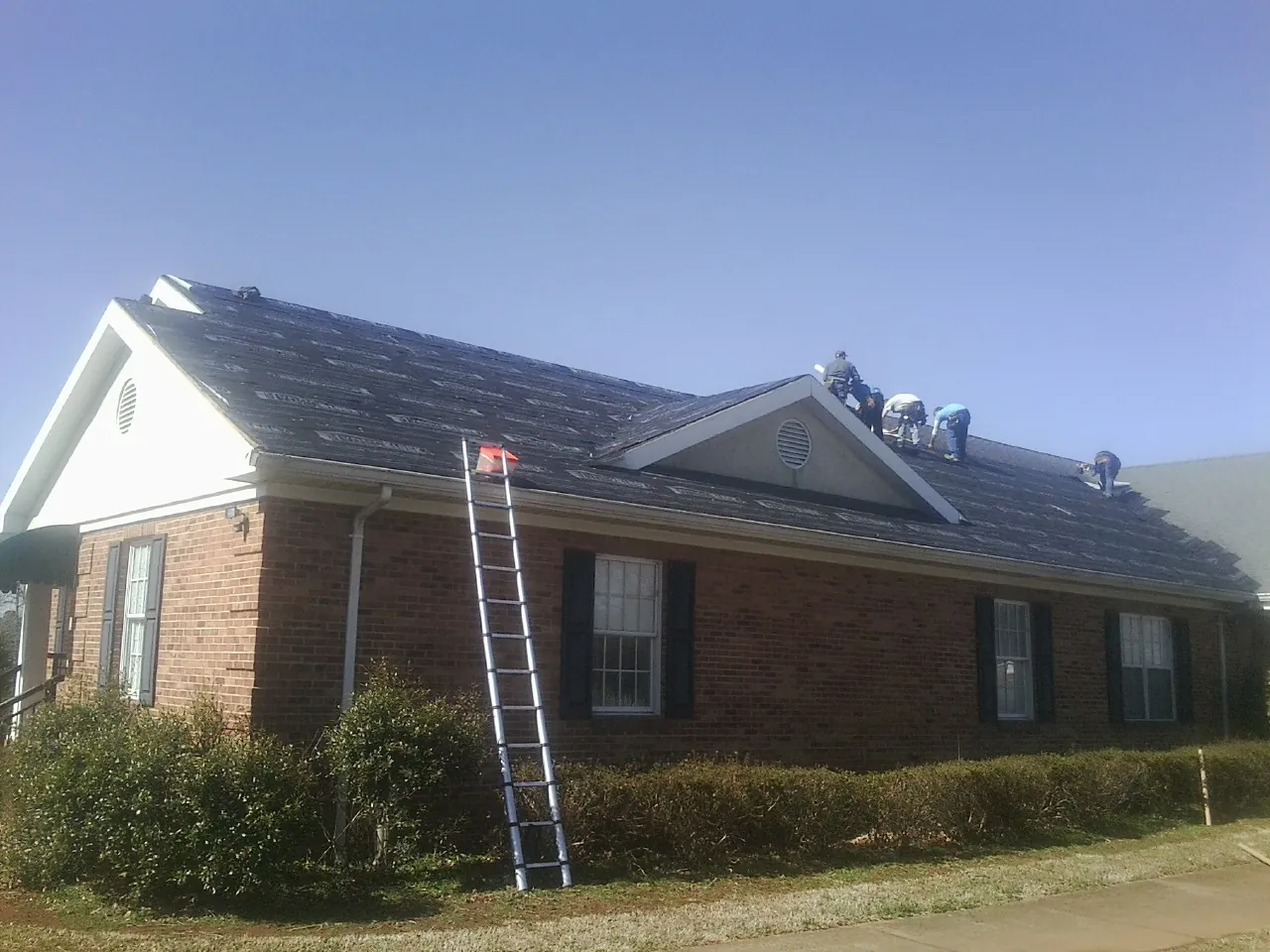 Crew tearing shingles off a brick ranch roof with a ladder set — Metal Roof Repair work by our crew in Decker